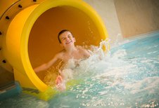 Junge mit strahlendem Lächeln rutscht aus einer gelben Wasserrutsche ins Becken des Landguts Furtherwirt und sorgt für spritzigen Badespaß