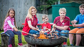 Drei Kinder gemeinsam mit zwei Kinderbetreuerinnen beim Lagerfeuer im Familienhotel Landgut Furtherwirt. Die Kinder grillen Stockbrot.