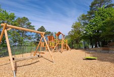 Spielplatz mit Klettergerüst, Nestschaukel und Karussell aus Holz im Familienhotel Sonnenhügel in der Rhön, umgeben von Bäumen und unter blauem Himmel.