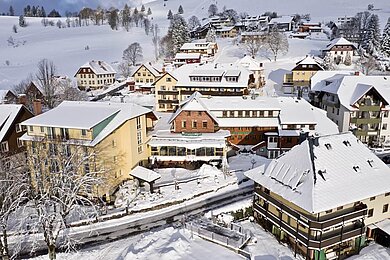 Luftaufnahme einer verschneiten Hotelanlage im Schwarzwald, umgeben von malerischen Häusern und schneebedeckten Bäumen, unterstrichen durch die idyllische Winterstimmung.