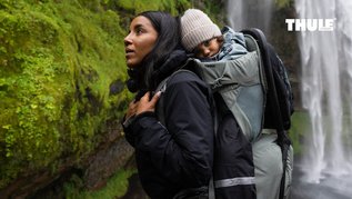 Frau wandert mit einem kleinen Kind in der Thule Kindertrage durch moosbedeckte Felslandschaft neben einem Wasserfall.