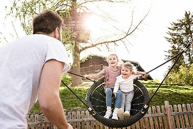 Vater mit zwei Kindern im Last Minute Familienurlaub im Familienhotel. Vater schaukelt die Mädchen auf dem Spielplatz in der Nestschaukel.