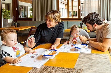 Eine Familie sitzt am Essenstisch und die Eltern füttern die beiden Babys im Familienhotel Das Bayrischzell in Oberbayern