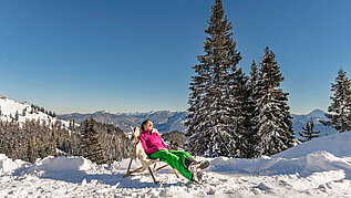 Eine Frau sitzt im Stuhl geniest die Sonne. Der Wald ist Schnee bedeckt.