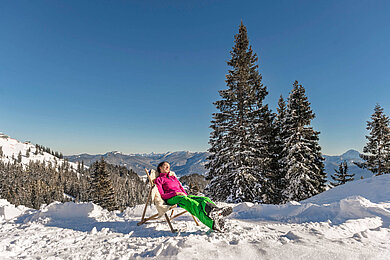 Eine Frau sitzt im Stuhl geniest die Sonne. Der Wald ist Schnee bedeckt.