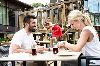 Familie beim Essen auf der Terrasse eines Hotels, währen die beiden Kinder auf einem Spielturm mit Eimer und Sand spielen.