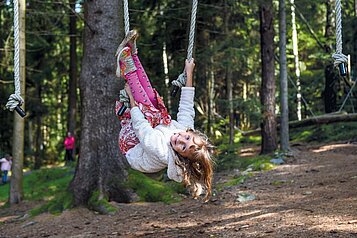 Kind hängt kopfüber an einem Seil im Waldspielplatz im Familienhotel Ulrichshof im Bayerischen Wald