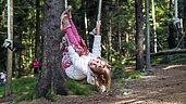 Kind hängt kopfüber an einem Seil im Waldspielplatz im Familienhotel Ulrichshof im Bayerischen Wald