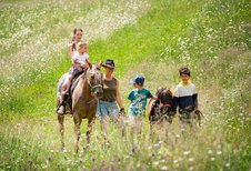 Kinder erleben gemeinsam mit einer Begleiterin einen Ponyritt über eine blühende Almwiese beim Familienhotel Bella Vista Südtirol.