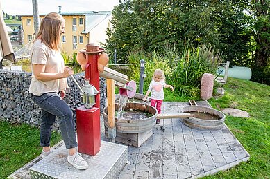 Eine Mutter mit ihrem Kinder auf einem Wasserspielplatz mit einer Wasserkanone im Garten des Familienhotel Engel im Schwarzwald.