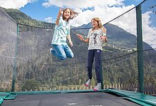 Zwei Mädchen springen auf dem Trampolin draußen auf dem Außengelände des Kinderhotels Sailer in Tirol.