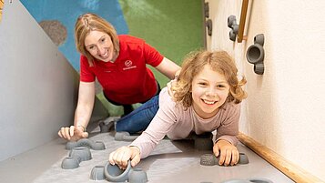 Mädchen klettert die Boulderwand im Happy-Club des Familienhotels Sonnenpark nach oben. Hinter ihr steht eine Kinderbetreuerin und passt auf sie auf.