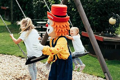 Maskottchen Happy schaukelt Kinder auf dem Outdoor-Spielplatz des Familienhotels Tirolerho