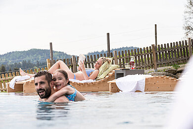 Familien Outdoorpool, Papa und tochter plantschen im Wasser. Mama liegt auf der Sonnenliege und liest ein Buch.