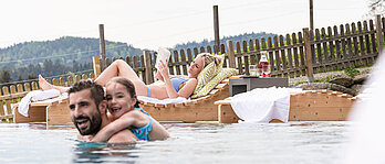 Familien Outdoorpool, Papa und tochter plantschen im Wasser. Mama liegt auf der Sonnenliege und liest ein Buch.