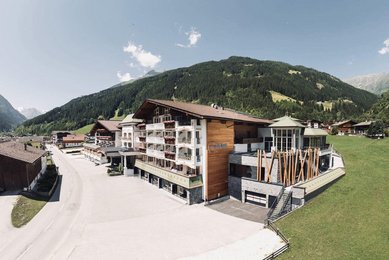 Sommeransicht des Familienhotels Kindl in den Tiroler Alpen mit umliegenden Wiesen, Bergen und traditioneller Architektur.