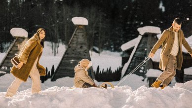 Familie mit Kind beim Winterspaziergang nahe des Alpura Retreats durch den Schnee, das Kind sitzt auf einem Holzschlitten, der von einem Erwachsenen gezogen wird.