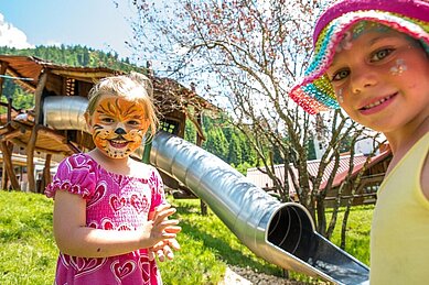 Zwei Mädchen sind im Gesicht bemalt und stehen vor einer Rutsche auf dem Spielplatz des Familienhotel Spa- & Familien-Resorts Krone im Allgäu ausklingen.