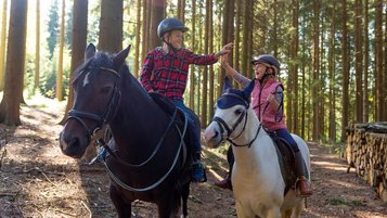 Junge und Mädchen reiten auf Pferden durch den Wald in der Nähe des Familienhotels Ulrichshof und geben sich ein glückliches High Five