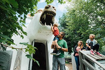 Familie ist auf dem Spielplatz und der Vater hebt sein Kind zum Bärenmaul hinauf.