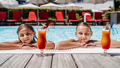 Zwei Mädchen genießen bunte Kinder-Cocktails am Poolrand des Familienhotels Amiamo an einem sonnigen Sommertag.
