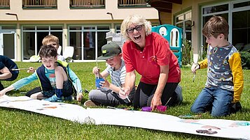 Kinder bemalen gemeinsam mit einer Kinderbetreuerin ein großes Plakat draußen in der Wiese des Familienhotels Lärchenhof in Tirol.