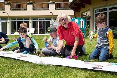 Kinder bemalen gemeinsam mit einer Kinderbetreuerin ein großes Plakat draußen in der Wiese des Familienhotels Lärchenhof in Tirol.