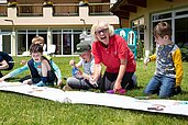 Kinder bemalen gemeinsam mit einer Kinderbetreuerin ein großes Plakat draußen in der Wiese des Familienhotels Lärchenhof in Tirol.