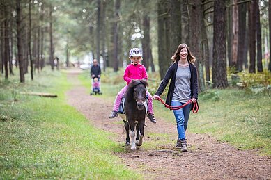 Eine Frau hält ein Pony an den Zügeln, auf dem ein Mädchen reitet.