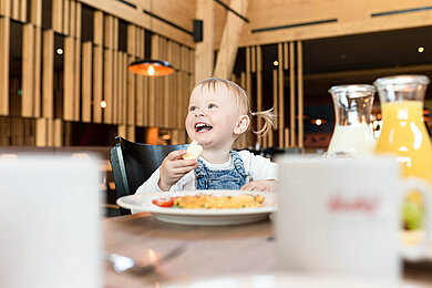 Ein Baby sitz am Tisch beim Essen und lacht und hat ein Stück Brot in der Hand.