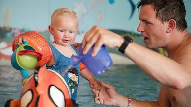 Ein blondes Kleinkind in blauer Badebekleidung spielt gemeinsam mit seinem Vater im warmen Babybecken des Familienhotels Huber, beide gießen Wasser über ein buntes Spielzeug im Wasser.