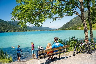 Familie macht eine Pause vom Fahrradfahren am See. Die Eltern sitzen auf einer Bank und die Kinder spielen am Wasser.