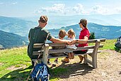Eltern genießen gemeinsam mit ihren Kindern auf einer Bank den schönen Ausblick in die Kärntner Berg