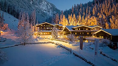 Das Hotel Habachklause im Salzburger Land erstrahlt bei Nacht in festlicher Beleuchtung, eingebettet in eine idyllische, schneebedeckte Landschaft mit dichten Tannen und einem beeindruckenden Berg im Hintergrund.