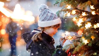 Kind schmückt einen beleuchteten Weihnachtsbaum im Schnee, warm eingepackt in Winterjacke und Strickmütze.