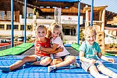 Drei Kinder sitzen auf dem Trampolin auf dem Außengelände des Familienhotels Das Hopfgarten.