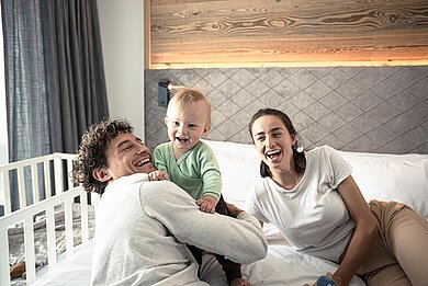 Baby sitzt lachend mit seinen Eltern auf dem Bett im Zimmer eines Familienhotels 