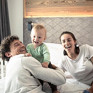 Baby sitzt lachend mit seinen Eltern auf dem Bett im Zimmer eines Familienhotels 