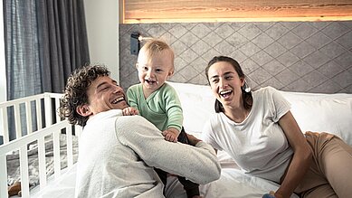 Baby sitzt lachend mit seinen Eltern auf dem Bett im Zimmer eines Familienhotels 