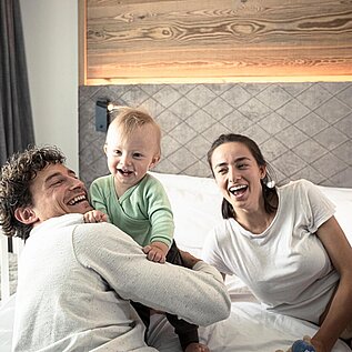 Baby sitzt lachend mit seinen Eltern auf dem Bett im Zimmer eines Familienhotels 