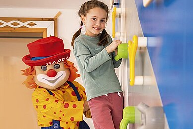 Kinder klettern an der Kletterwand im Happy-Club des Familienhotels Alpenhotel Kindl in Tirol. Auch Clown Happy, das Maskottchen des Familienhotels, klettert mit und bringt die Kinder zum Lachen.