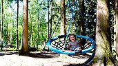 Ein Kind lacht uns sitzt in einer Nestschaukel im Waldspielplatz im Familienhotel Ulrichshof im Bayerischen Wald