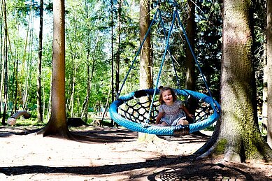 Ein Kind lacht uns sitzt in einer Nestschaukel im Waldspielplatz im Familienhotel Ulrichshof im Bayerischen Wald