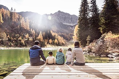 Eine Familie mit drei Kindern sitzt im Herbst auf einem Holzsteg und blickt auf den See und die Berge.