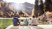 Eine Familie mit drei Kindern sitzt im Herbst auf einem Holzsteg und blickt auf den See und die Berge.