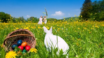 Weißer Hase neben einem Korb mit bunten Ostereiern auf einer blühenden Wiese, im Hintergrund zwei Kinder unter blauem Himmel.