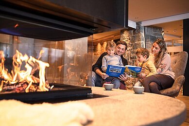 Eltern sitzen mit ihren zwei Kindern vor einem Kaminfeuer und lesen den Kindern aus einem Buch vor.