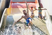 Ein Vater rutscht mit seinen beiden Kindern die Wasserrutsche in den Außenpool im Family Home Alpenhof Familienhotel in Südtirol. 