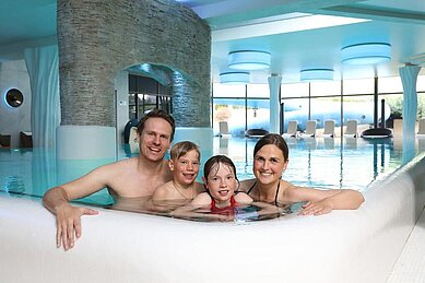 Zwei fröhliche Kinder spielen mit Wasserattraktionen in einem Kinderbeckenbereich des Hotels Feldberger Hof, spritzendes Wasser und leuchtende Farben im Hintergrund.