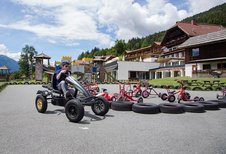Ein Kind fährt mit einem Gokart auf einer Outdoor-Bahn, umgeben von Spielgeräten und Tricycles, mit dem Kärntner Familienhotel Ramsi im Hintergrund